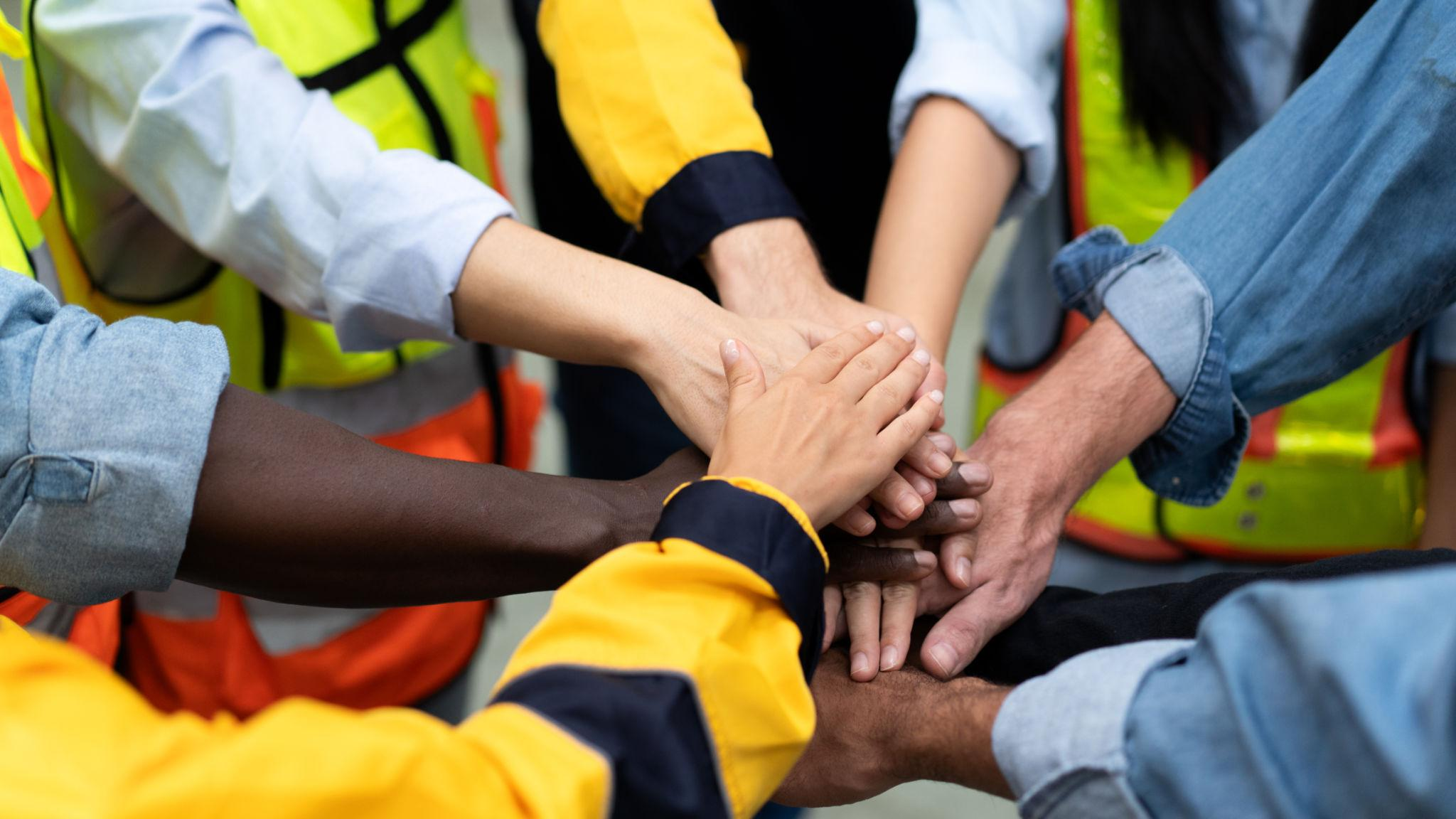 Team members in safety vests stacking hands together in a circle, symbolizing collaboration and unity.