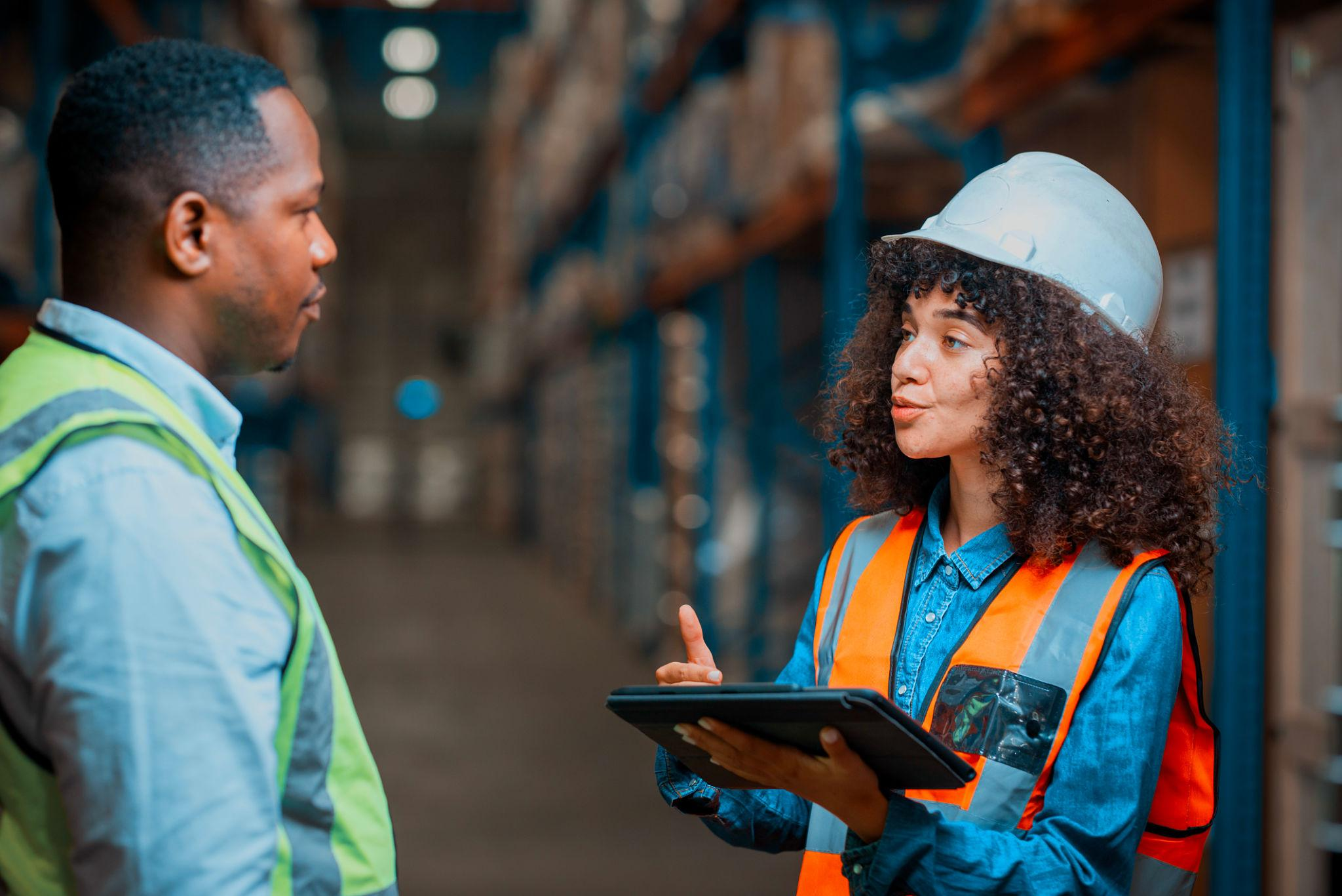 Two logistics professionals in safety vests discussing operations with a tablet in a warehouse aisle.
